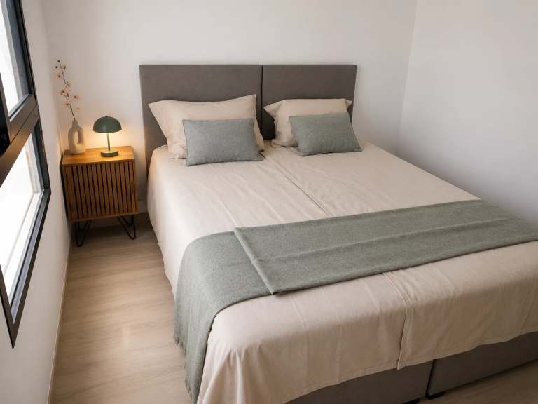 Lichte slaapkamer met comfortabel tweepersoonsbed, grijs hoofdbord, houten nachtkastje en sfeervolle lamp.