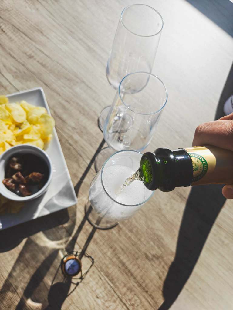 Hand vult champagneglas met bubbels, naast schaal met chips en nootjes op lichte houten tafel in de zon